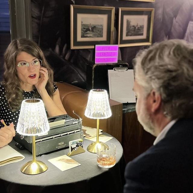 A typewriter poet talks with a guest across a lamplit table before typing his poem