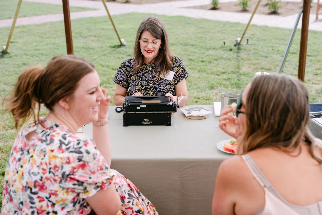 Laura Frantz typing at her Royal typewriter under an outdoor tent while two guests wait at the table