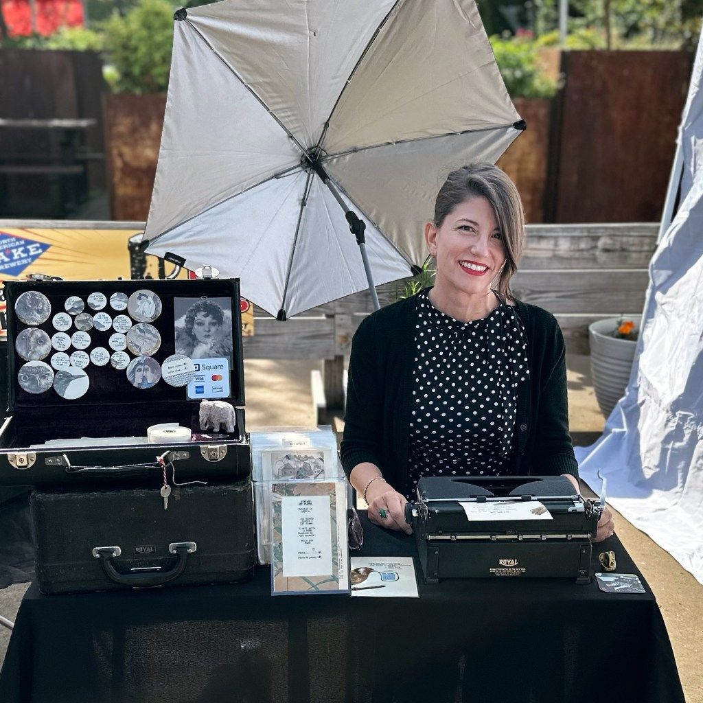 Laura Frantz smiling behind her typewriter table at an outdoor market